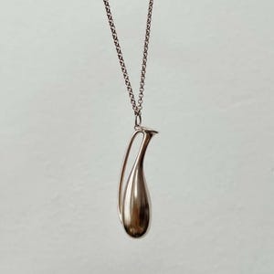 May include: A silver-toned pendant necklace featuring a teardrop-shaped charm with a curved handle. The charm is smooth and reflective, suspended from a delicate chain. The necklace is displayed against a plain, light background.