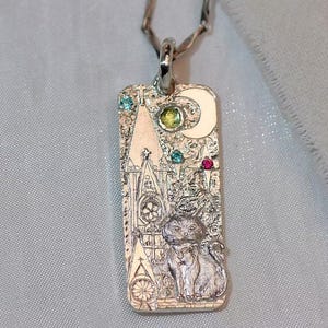 May include: Silver pendant necklace with a rectangular charm featuring a cat, a crescent moon, and a cathedral design. The charm is adorned with colorful gemstones and hangs from a twisted silver chain.