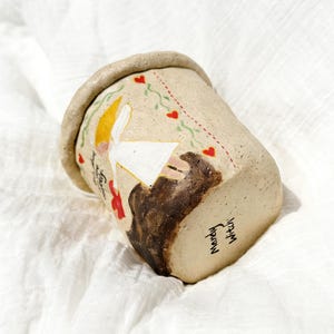 Puede incluir: Una taza de cer&aacute;mica hecha a mano con un exterior beige, que presenta una ilustraci&oacute;n caprichosa de una figura con cabeza amarilla y vestido blanco, y un animal marr&oacute;n. La taza tiene un borde marr&oacute;n y est&aacute; decorada con corazones rojos y vides verdes. El texto "Mandy Witch" est&aacute; escrito en la parte inferior.