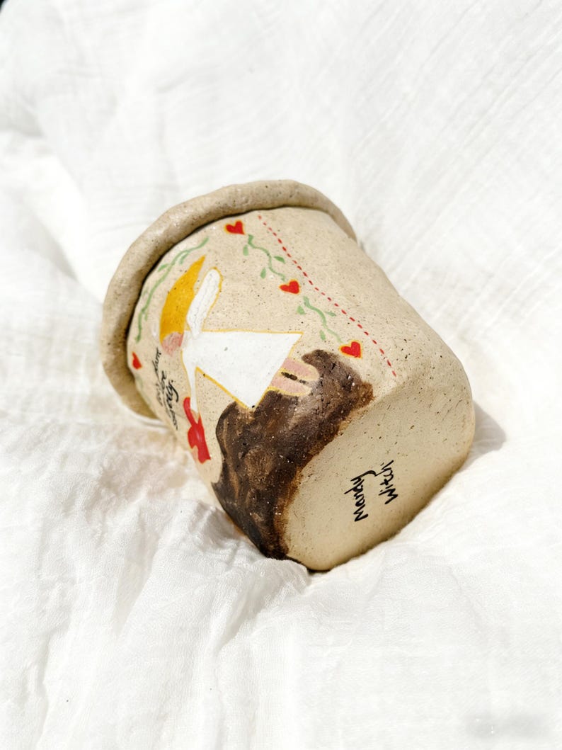 Puede incluir: Una taza de cer&aacute;mica hecha a mano con un exterior beige, que presenta una ilustraci&oacute;n caprichosa de una figura con cabeza amarilla y vestido blanco, y un animal marr&oacute;n. La taza tiene un borde marr&oacute;n y est&aacute; decorada con corazones rojos y vides verdes. El texto "Mandy Witch" est&aacute; escrito en la parte inferior.