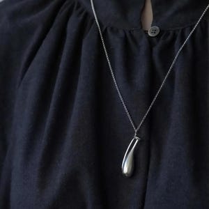 May include: A silver pendant necklace with a teardrop-shaped charm. The charm is smooth and reflective, hanging from a delicate chain. The necklace is displayed against a dark navy blue fabric.