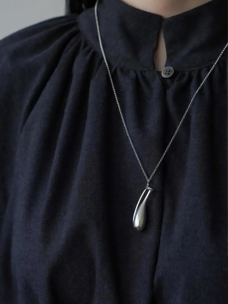 May include: A silver pendant necklace with a teardrop-shaped charm. The charm is smooth and reflective, hanging from a delicate chain. The necklace is displayed against a dark navy blue fabric.