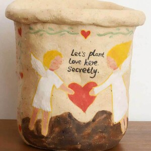 Puede incluir: Una maceta de cer&aacute;mica beige con borde enrollado, con un dise&ntilde;o pintado a mano. El dise&ntilde;o incluye dos &aacute;ngeles, un coraz&oacute;n rojo y el texto "Let's plant love here. Secretly."