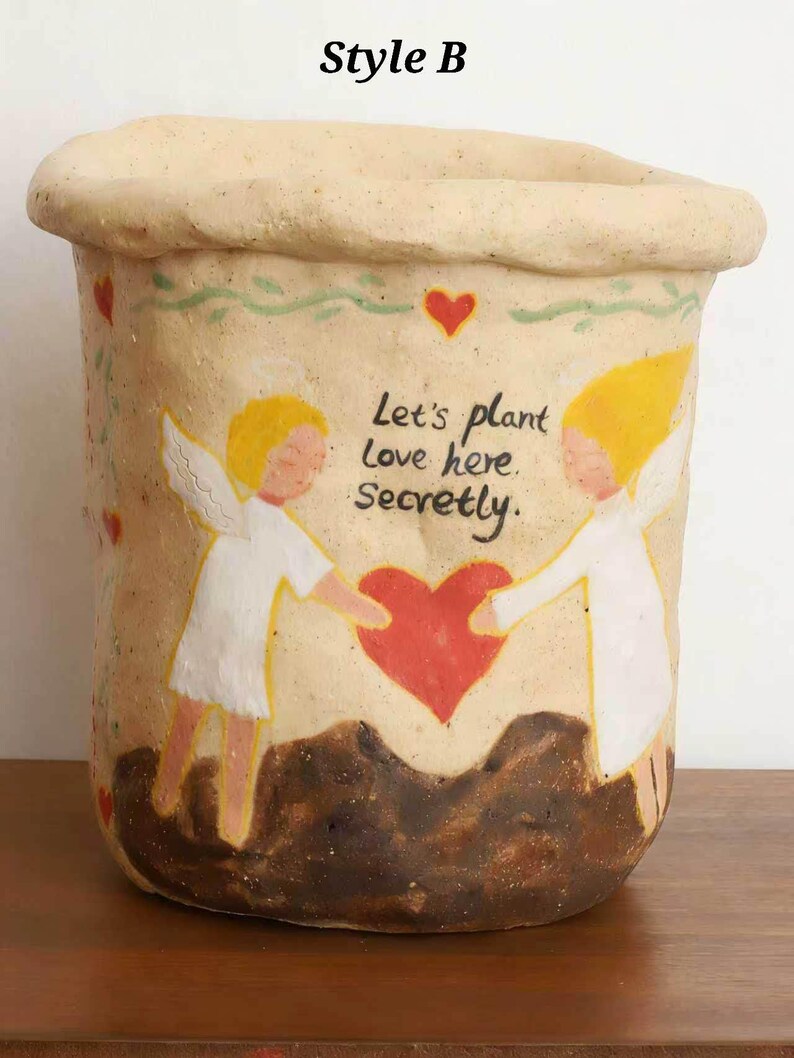 Puede incluir: Una maceta de cer&aacute;mica beige con borde enrollado, con un dise&ntilde;o pintado a mano. El dise&ntilde;o incluye dos &aacute;ngeles, un coraz&oacute;n rojo y el texto "Let's plant love here. Secretly."