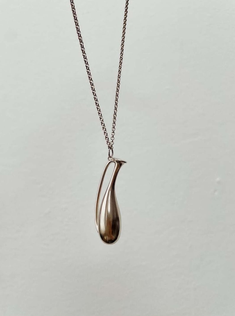 May include: A silver-toned pendant necklace featuring a teardrop-shaped charm with a curved handle. The charm is smooth and reflective, suspended from a delicate chain. The necklace is displayed against a plain, light background.