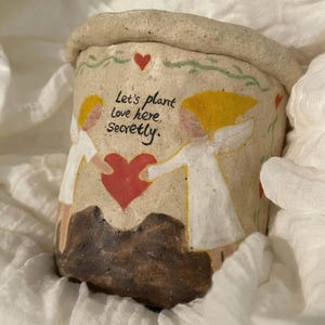 Puede incluir: Una maceta de cer&aacute;mica beige con borde enrollado. La maceta presenta un dise&ntilde;o pintado de dos figuras sosteniendo un coraz&oacute;n rojo, con el texto "Let's plant Love here, Secretly." Tambi&eacute;n hay corazones y vides decorativas pintadas en la maceta.