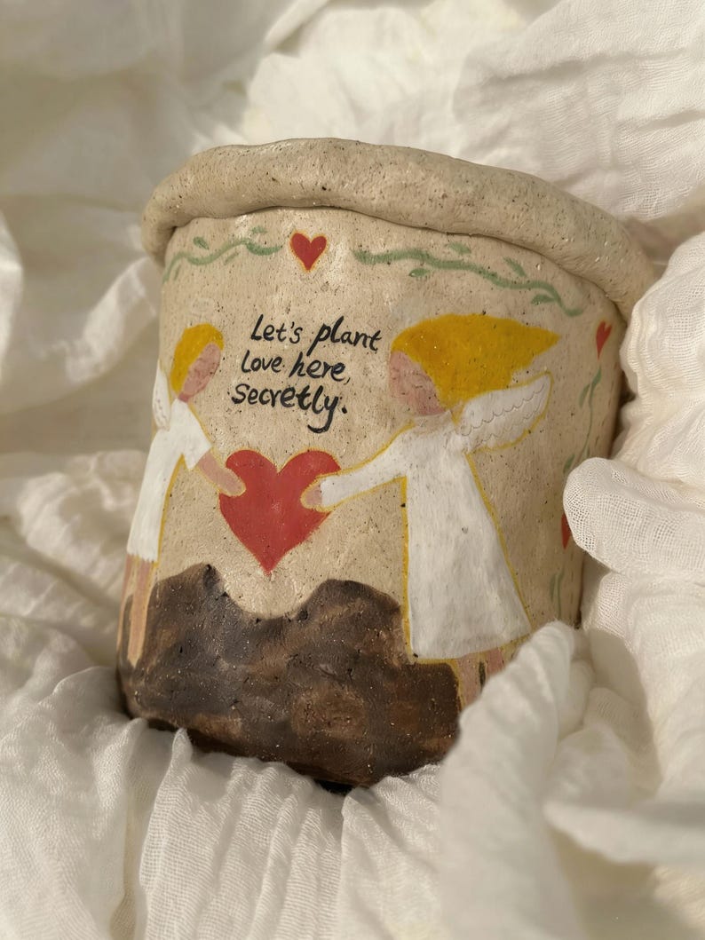 Puede incluir: Una maceta de cer&aacute;mica beige con borde enrollado. La maceta presenta un dise&ntilde;o pintado de dos figuras sosteniendo un coraz&oacute;n rojo, con el texto "Let's plant Love here, Secretly." Tambi&eacute;n hay corazones y vides decorativas pintadas en la maceta.