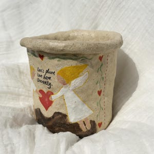 Puede incluir: Una peque&ntilde;a maceta de cer&aacute;mica beige con borde enrollado. La maceta presenta un dise&ntilde;o pintado a mano de dos &aacute;ngeles sosteniendo un coraz&oacute;n rojo, con el texto "Let's plant love here secretly". Corazones rojos y vides verdes decoran los lados.