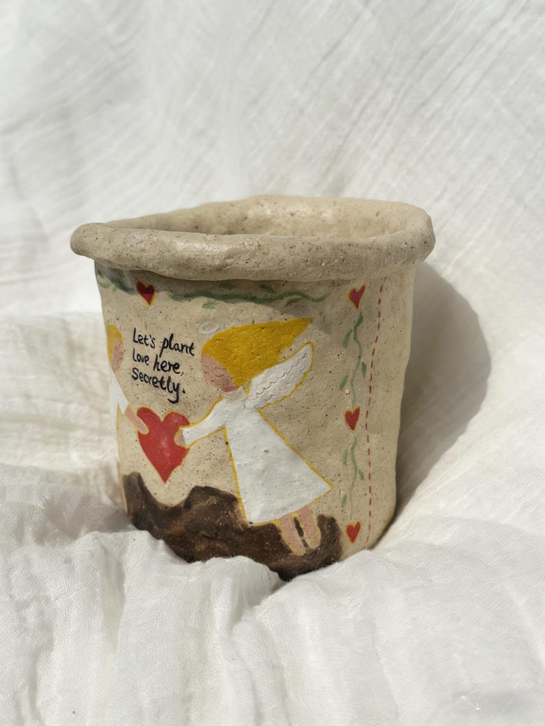 Puede incluir: Una peque&ntilde;a maceta de cer&aacute;mica beige con borde enrollado. La maceta presenta un dise&ntilde;o pintado a mano de dos &aacute;ngeles sosteniendo un coraz&oacute;n rojo, con el texto "Let's plant love here secretly". Corazones rojos y vides verdes decoran los lados.