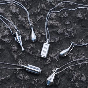 May include: A collection of six silver-toned metal pendants on delicate matching chains. The pendants feature various geometric and vessel-like shapes, including cylindrical, teardrop, and amphora-inspired designs with smooth, reflective surfaces.