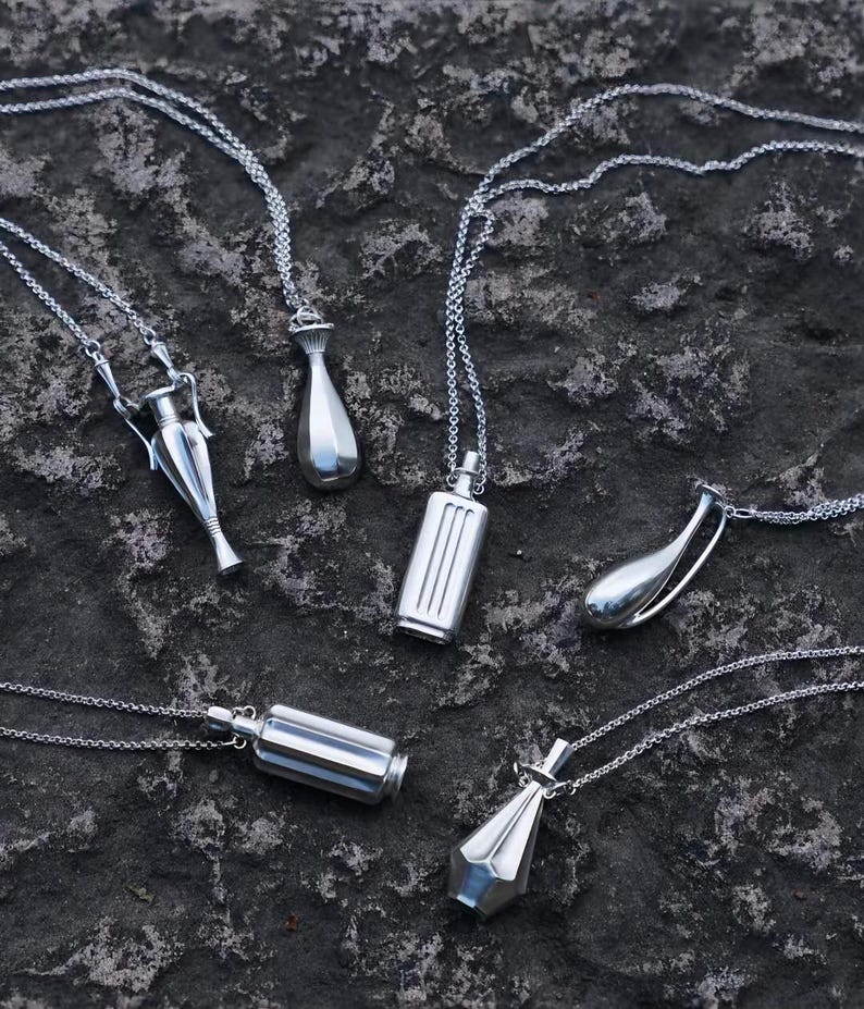 May include: A collection of six silver-toned metal pendants on delicate matching chains. The pendants feature various geometric and vessel-like shapes, including cylindrical, teardrop, and amphora-inspired designs with smooth, reflective surfaces.