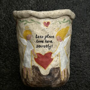 Puede incluir: Maceta de cer&aacute;mica beige con superficie texturizada. Presenta dos &aacute;ngeles con cabello amarillo y t&uacute;nicas blancas, sosteniendo un coraz&oacute;n rojo. El texto "Let's plant love here, secretly!" est&aacute; escrito en la maceta.