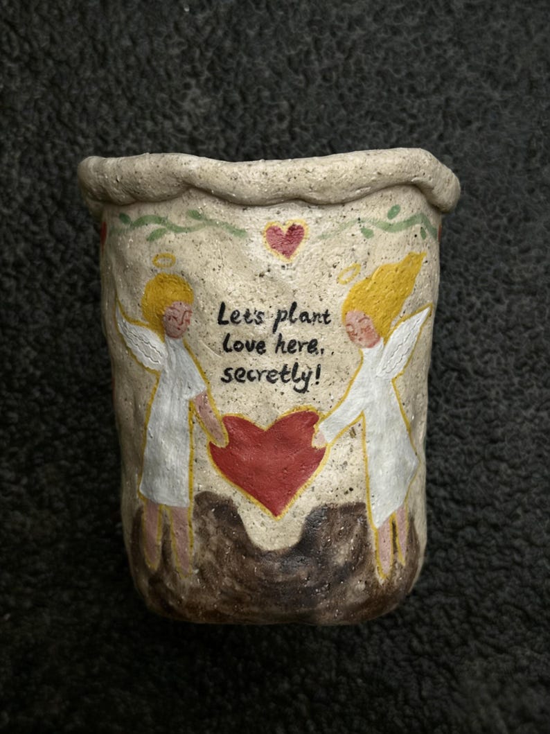 Puede incluir: Maceta de cer&aacute;mica beige con superficie texturizada. Presenta dos &aacute;ngeles con cabello amarillo y t&uacute;nicas blancas, sosteniendo un coraz&oacute;n rojo. El texto "Let's plant love here, secretly!" est&aacute; escrito en la maceta.
