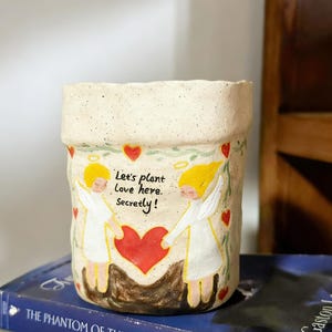 Puede incluir: Una maceta de cer&aacute;mica color crema con una superficie texturizada. La maceta presenta un dise&ntilde;o pintado de dos &aacute;ngeles sosteniendo un coraz&oacute;n rojo, con el texto "Let's plant love here, secretly!" rodeado de corazones rojos y vides verdes.
