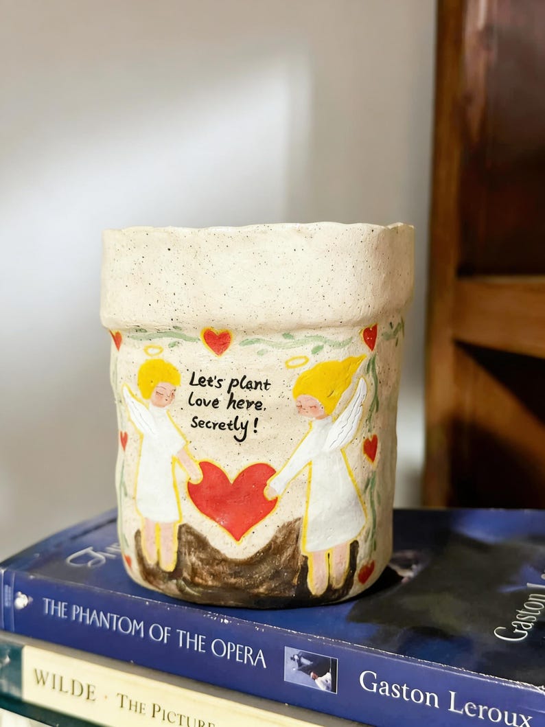 Puede incluir: Una maceta de cer&aacute;mica color crema con una superficie texturizada. La maceta presenta un dise&ntilde;o pintado de dos &aacute;ngeles sosteniendo un coraz&oacute;n rojo, con el texto "Let's plant love here, secretly!" rodeado de corazones rojos y vides verdes.