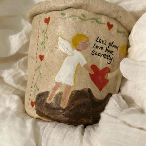 Puede incluir: Una maceta de cer&aacute;mica beige con borde enrollado. La maceta est&aacute; decorada con corazones rojos, vides verdes y el texto "Let's plant Love here. Secretly." Dos figuras de &aacute;ngeles est&aacute;n pintadas en el frente, sosteniendo un coraz&oacute;n rojo.