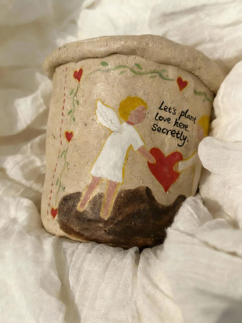 Puede incluir: Una maceta de cer&aacute;mica beige con borde enrollado. La maceta est&aacute; decorada con corazones rojos, vides verdes y el texto "Let's plant Love here. Secretly." Dos figuras de &aacute;ngeles est&aacute;n pintadas en el frente, sosteniendo un coraz&oacute;n rojo.