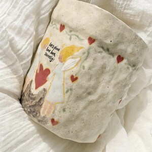 Puede incluir: Una maceta de cer&aacute;mica hecha a mano con un acabado texturizado en blanco roto. La maceta presenta una ilustraci&oacute;n caprichosa de dos figuras con alas de &aacute;ngel, corazones rojos y el texto "Let's plant love here, surely!". En la superficie est&aacute;n pintadas vides decorativas y m&aacute;s corazones.