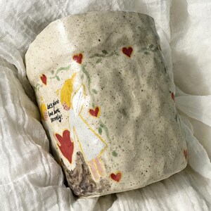 Puede incluir: Taza de cer&aacute;mica hecha a mano con una superficie texturizada y moteada. La taza presenta un dise&ntilde;o caprichoso de una figura con alas de &aacute;ngel, corazones rojos y el texto "Let's plant love here, surely!" en un estilo manuscrito. La taza est&aacute; sobre una tela blanca.