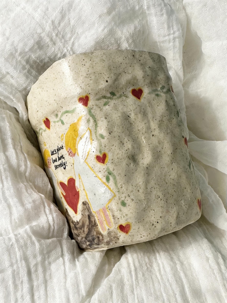 Puede incluir: Taza de cer&aacute;mica hecha a mano con una superficie texturizada y moteada. La taza presenta un dise&ntilde;o caprichoso de una figura con alas de &aacute;ngel, corazones rojos y el texto "Let's plant love here, surely!" en un estilo manuscrito. La taza est&aacute; sobre una tela blanca.