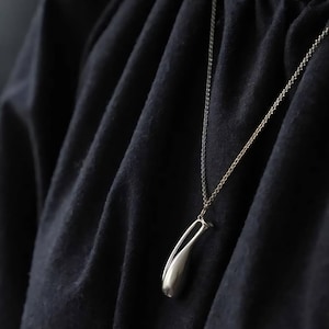 May include: A silver-toned pendant shaped like a stylized, elongated teardrop or bud, suspended from a delicate matching chain. The pendant features a smooth, polished metallic finish and is worn against a dark, textured fabric garment.