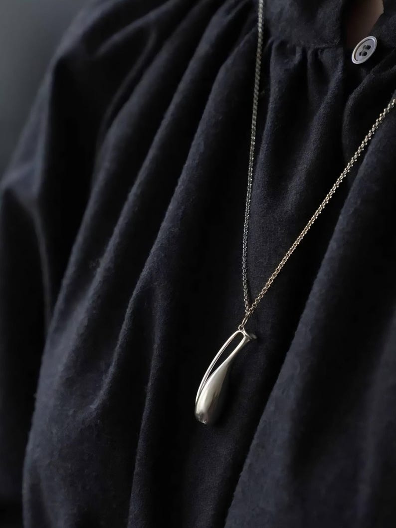 May include: A silver-toned pendant shaped like a stylized, elongated teardrop or bud, suspended from a delicate matching chain. The pendant features a smooth, polished metallic finish and is worn against a dark, textured fabric garment.
