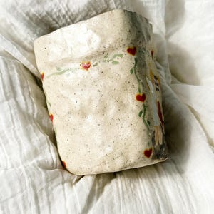 Puede incluir: Una taza de cer&aacute;mica hecha a mano con una superficie texturizada y moteada. La taza blanquecina presenta un dise&ntilde;o de corazones rojos y vides verdes. Tambi&eacute;n es visible una ilustraci&oacute;n caprichosa de una figura.