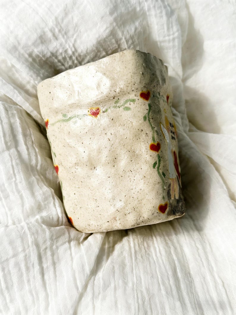 Puede incluir: Una taza de cer&aacute;mica hecha a mano con una superficie texturizada y moteada. La taza blanquecina presenta un dise&ntilde;o de corazones rojos y vides verdes. Tambi&eacute;n es visible una ilustraci&oacute;n caprichosa de una figura.