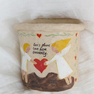 Puede incluir: Una maceta de cer&aacute;mica beige con borde enrollado. La maceta presenta un dise&ntilde;o pintado de dos &aacute;ngeles sosteniendo un coraz&oacute;n rojo, con el texto "Let's plant love here, secretly". Corazones rojos y vides verdes decoran el borde.