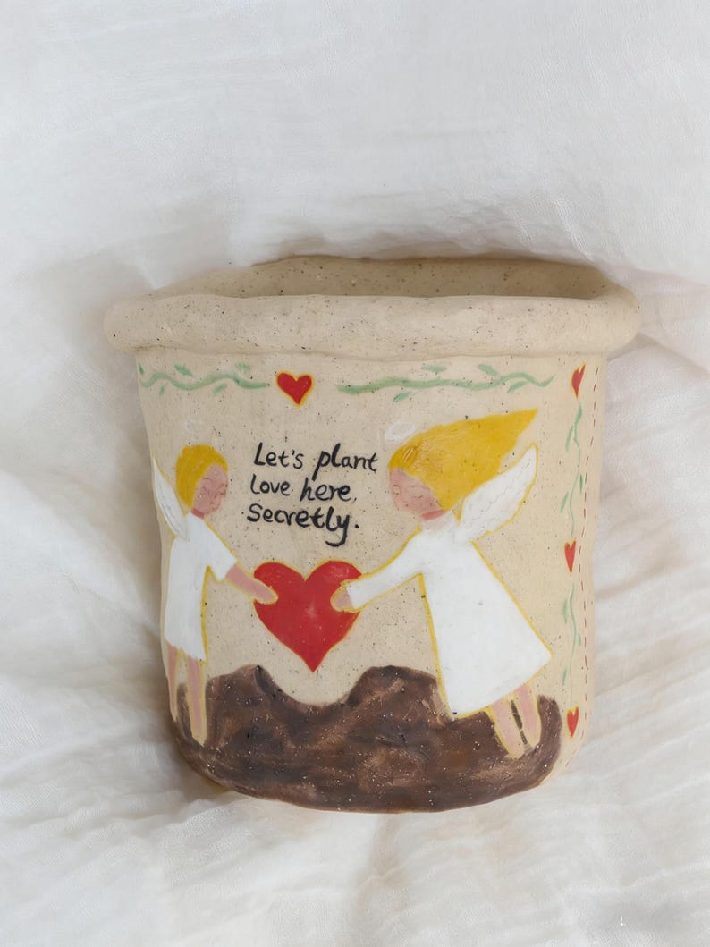 Puede incluir: Una maceta de cer&aacute;mica beige con borde enrollado. La maceta presenta un dise&ntilde;o pintado de dos &aacute;ngeles sosteniendo un coraz&oacute;n rojo, con el texto "Let's plant love here, secretly". Corazones rojos y vides verdes decoran el borde.