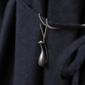 May include: A silver-colored pendant necklace featuring a miniature pitcher design. The pendant hangs from a delicate chain against a dark blue fabric background. The pitcher has a smooth, polished surface and a curved handle.