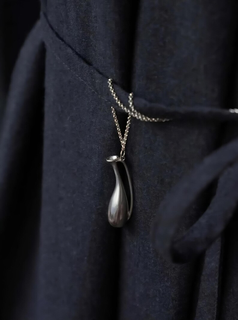 May include: A silver-colored pendant necklace featuring a miniature pitcher design. The pendant hangs from a delicate chain against a dark blue fabric background. The pitcher has a smooth, polished surface and a curved handle.