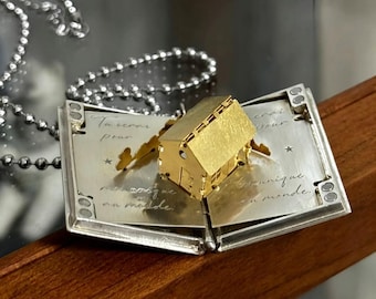 May include: A silver-toned metal locket shaped like an open book with a gold-toned three-dimensional house pop-up inside. The interior pages feature engraved French cursive text and small star accents. A silver ball chain is attached to the locket.