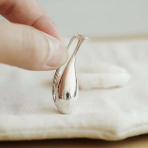 May include: A small, polished silver pendant shaped like a slender, elongated amphora or vase. The metallic surface is highly reflective, featuring a smooth, curved body with a narrow neck and a single delicate handle attached at the top.
