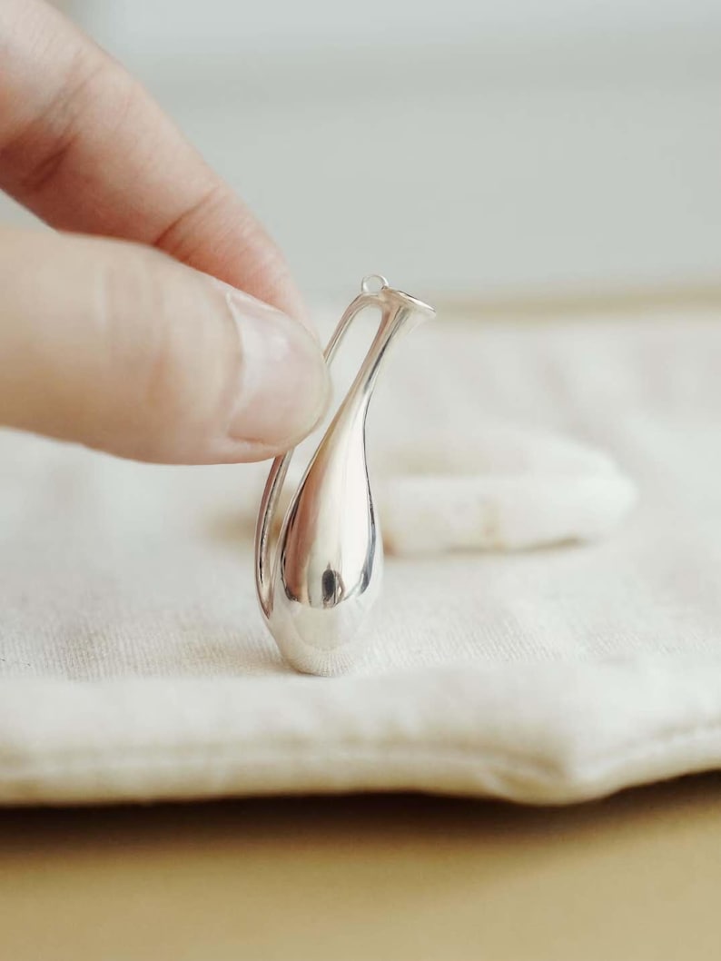 May include: A small, polished silver pendant shaped like a slender, elongated amphora or vase. The metallic surface is highly reflective, featuring a smooth, curved body with a narrow neck and a single delicate handle attached at the top.