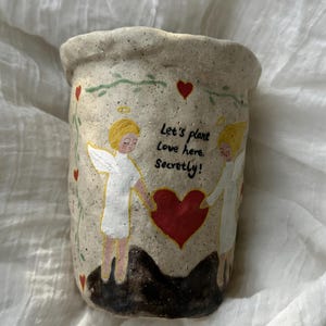 Puede incluir: Una maceta de cer&aacute;mica beige con superficie texturizada. La maceta presenta un dise&ntilde;o pintado de dos &aacute;ngeles sosteniendo un coraz&oacute;n rojo, con el texto "Let's plant Love here. Secretly!" y detalles de corazones rojos.