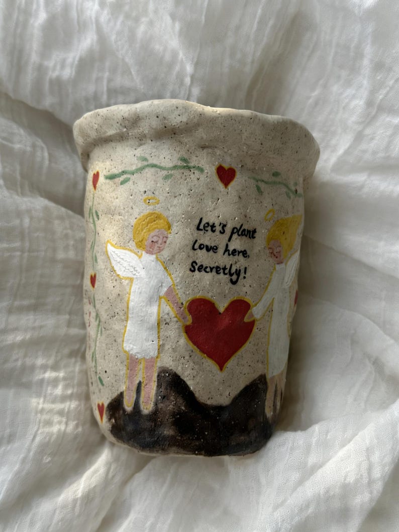 Puede incluir: Una maceta de cer&aacute;mica beige con superficie texturizada. La maceta presenta un dise&ntilde;o pintado de dos &aacute;ngeles sosteniendo un coraz&oacute;n rojo, con el texto "Let's plant Love here. Secretly!" y detalles de corazones rojos.