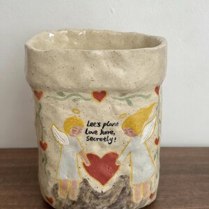 Puede incluir: Jardinera de cer&aacute;mica beige con superficie texturizada. La jardinera presenta un dise&ntilde;o de dos &aacute;ngeles tomados de la mano sobre un coraz&oacute;n rojo, con el texto "Let's plant Love here, Secretly!". Corazones rojos y vides verdes decoran la jardinera.
