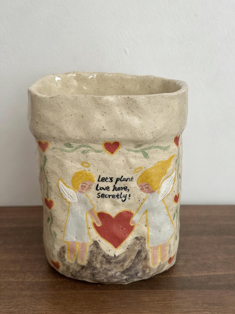Puede incluir: Jardinera de cer&aacute;mica beige con superficie texturizada. La jardinera presenta un dise&ntilde;o de dos &aacute;ngeles tomados de la mano sobre un coraz&oacute;n rojo, con el texto "Let's plant Love here, Secretly!". Corazones rojos y vides verdes decoran la jardinera.