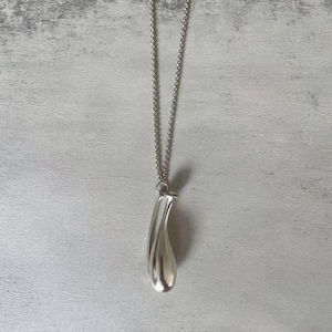 May include: A silver-toned necklace featuring a long, smooth, teardrop-shaped pendant suspended from a delicate, thin metal chain. The pendant has a polished, reflective surface with a curved, elongated form that tapers to a rounded base.