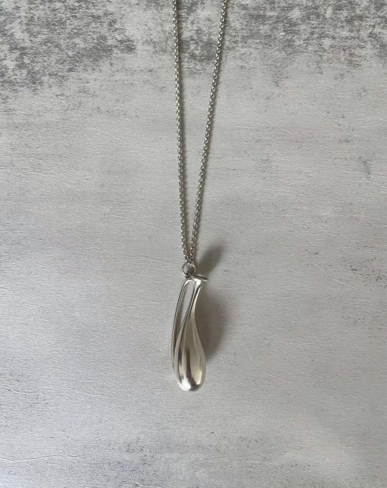 May include: A silver-toned necklace featuring a long, smooth, teardrop-shaped pendant suspended from a delicate, thin metal chain. The pendant has a polished, reflective surface with a curved, elongated form that tapers to a rounded base.