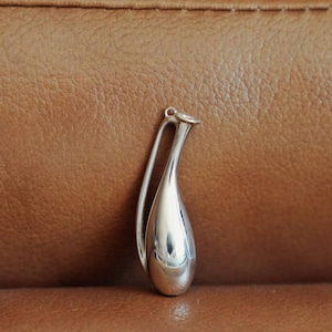 May include: A polished silver pendant in a teardrop shape with a curved handle. The pendant is smooth and reflective, catching the light. The design is minimalist and elegant, suitable for jewelry. The background is a textured brown surface.
