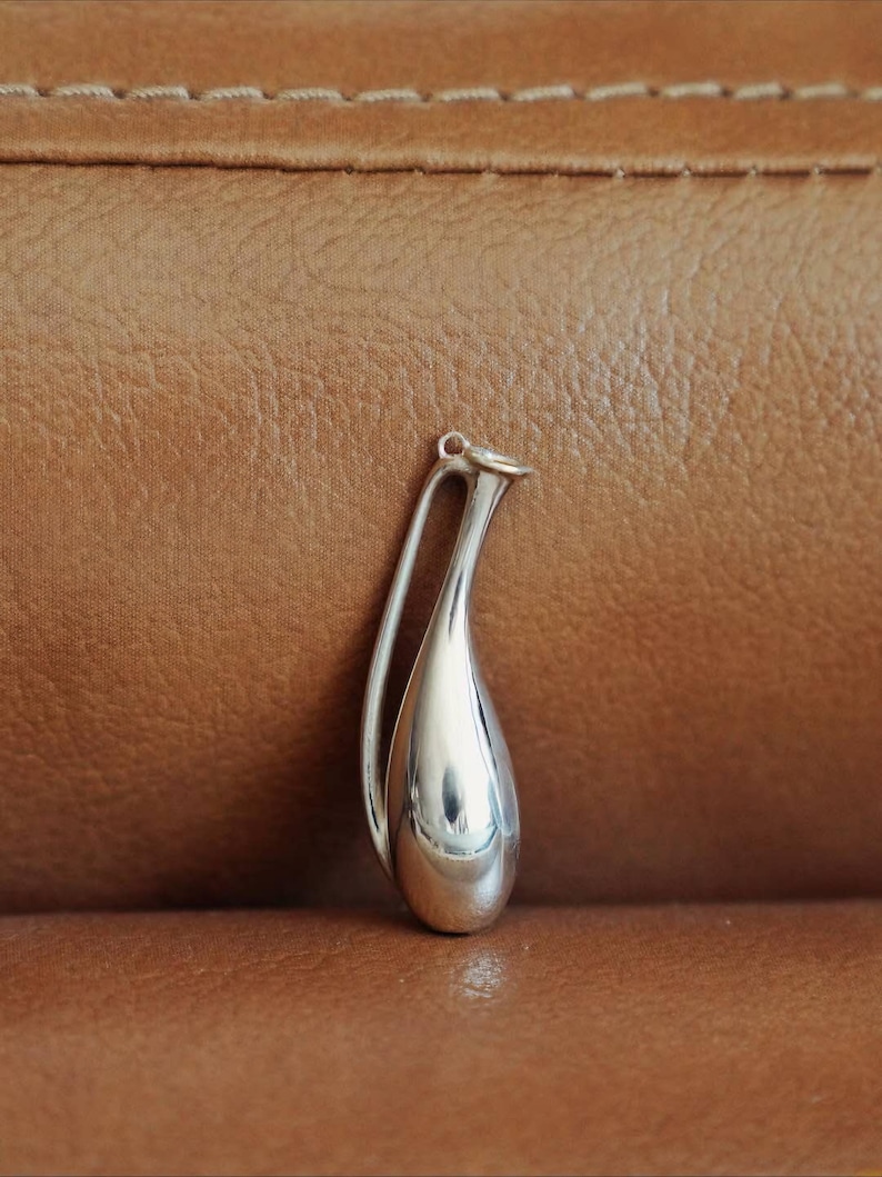 May include: A polished silver pendant in a teardrop shape with a curved handle. The pendant is smooth and reflective, catching the light. The design is minimalist and elegant, suitable for jewelry. The background is a textured brown surface.