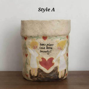 Puede incluir: Una maceta de cer&aacute;mica beige con superficie texturizada. La maceta presenta un dise&ntilde;o pintado de dos &aacute;ngeles sosteniendo un coraz&oacute;n rojo, con el texto "Let's plant Love here, secretly!" y detalles de corazones rojos.