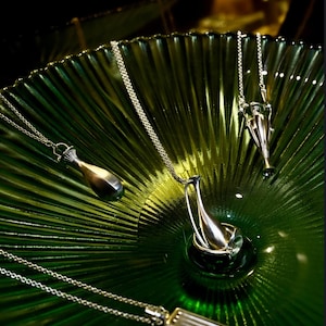 May include: Silver pendant necklaces with unique designs are displayed on a textured green glass surface. The pendants have various shapes, including a bottle, a vase, and a rectangular bar. The chains are delicate.