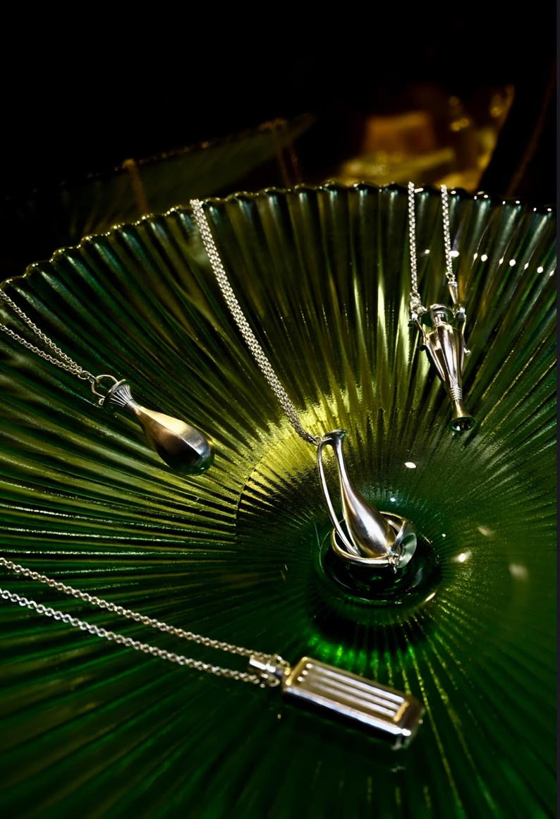 May include: Silver pendant necklaces with unique designs are displayed on a textured green glass surface. The pendants have various shapes, including a bottle, a vase, and a rectangular bar. The chains are delicate.