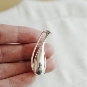 May include: A polished silver pendant shaped like a stylized teardrop or elongated vase. The piece features a smooth, reflective metallic surface with a hollow, open-work design at the top, terminating in a small circular loop for attachment.