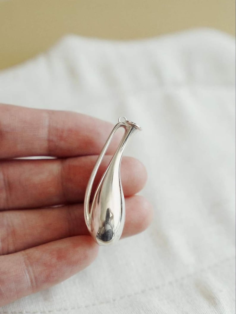 May include: A polished silver pendant shaped like a stylized teardrop or elongated vase. The piece features a smooth, reflective metallic surface with a hollow, open-work design at the top, terminating in a small circular loop for attachment.