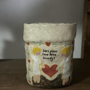Puede incluir: Una maceta de cer&aacute;mica hecha a mano con un acabado texturizado y moteado. La maceta presenta un dise&ntilde;o pintado de dos &aacute;ngeles sosteniendo un coraz&oacute;n, con el texto "Let's plant love here, secretly!" en negro. Corazones rojos est&aacute;n esparcidos alrededor de los &aacute;ngeles.