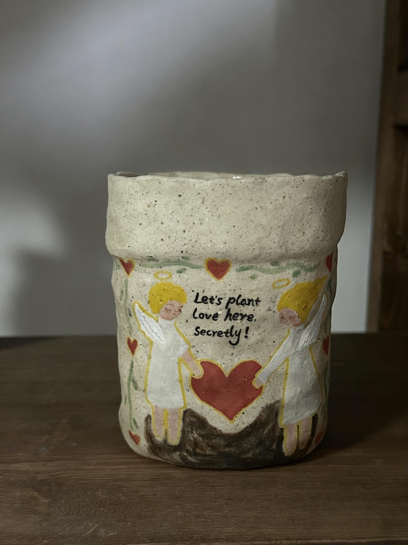 Puede incluir: Una maceta de cer&aacute;mica hecha a mano con un acabado texturizado y moteado. La maceta presenta un dise&ntilde;o pintado de dos &aacute;ngeles sosteniendo un coraz&oacute;n, con el texto "Let's plant love here, secretly!" en negro. Corazones rojos est&aacute;n esparcidos alrededor de los &aacute;ngeles.